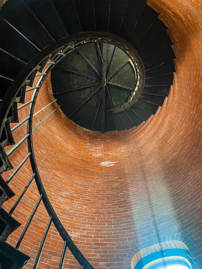 Looking up inside a lighthouse, this image shows a view of a black metal spiral staircase winding tightly against the curved red brick walls. The staircase forms a coil above, leading the eye upward toward the dark metal ceiling at the top. A metal railing runs alongside the stairs. At the bottom of the image, soft natural light enters through an arched window, casting a pale blue glow onto the bricks.