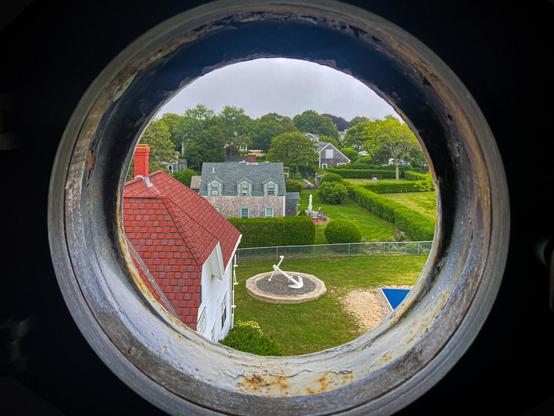 The photo is taken through a round, weathered porthole window from inside a lighthouse. The view looks out onto a tidy green yard with neatly trimmed hedges and several historic-style houses with gray shingles and red or gray roofs. In the center of the yard is a large white anchor mounted upright in a circular bed of gravel. Trees and shrubs fill the background, and the sky is cloudy and overcast.