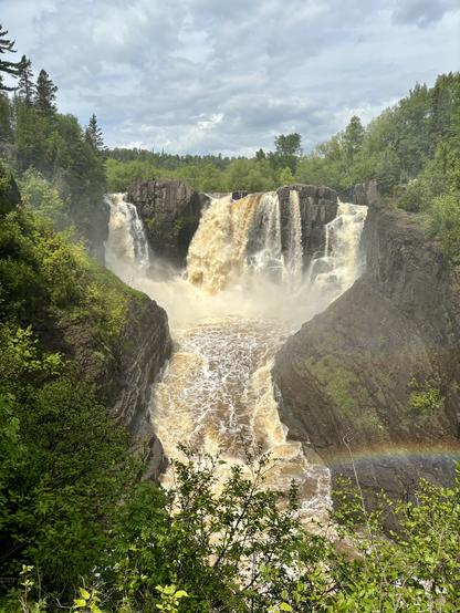 Looking up the Pigeon River High Falls. The right bank, Canada. The left bank, U.S. A main cascade in the center is creamy and slightly brown with high waters from inland. It is flanked by several smaller falls on either side. Water passes through a small gorge over which a rainbow is seen.