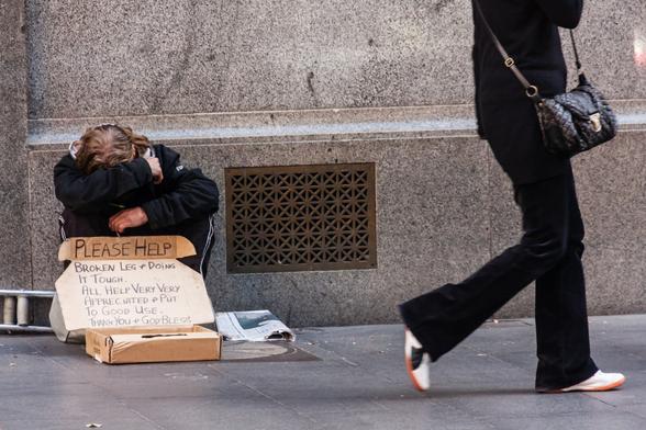 Against a stone wall of a city building, a man is crouching or possibly sitting on something, with his head downwards and face covered by his arm.  By the look of his hands, though, he may be quite young. In front of him, there's a box for collecting money and a sign that says:  Please help. Broken leg and doing it tough. All help very appreciated and put a good use. Thank you and God bless. To his right, a woman with a fashionable handbag, but only visible from shoulder level downwards, walks past.