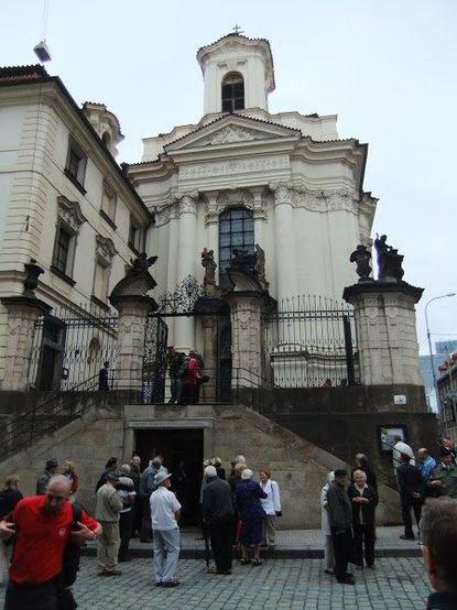 the Baroque church seen from across the street in front of the Paratroopers' Pub (Krčma u Parašutistů)

White Baroque structure with a central tower and a courtyard in front, built 1730-36
the view is of the church entrance above, inside an iron fence, and the entrance to the memorial and crypt, below it, beneath the stairs that ascend on both sides

the people assembled in front have just come from the streetside annual ceremony 

Although this was an Orthodox church, the Nazis persisted in calling it by its old name, Karel Boromäus--Counterreformation saint Carlo Borromeo
