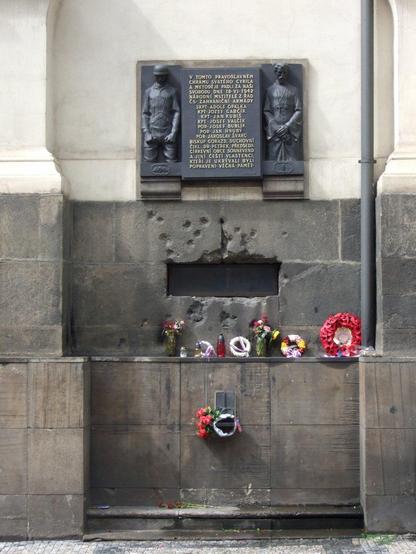 the bullet-pocked wall of the church:

a ventilation grate provided access to the crypt, and gunfire was exchanged from within and without. Eventually, the Nazis tried to drown the paratroopers by pumping water into the crypt through this opening

above the opening is a memorial plaque (next photo)

beneath it on a small ledge are small memorial wreaths and candles