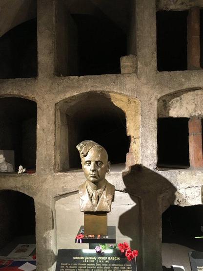 bronze bust of assassin Jozef Gabčík in garrison cap in front of the burial cubicles

beneath each bust is a black plaque with names and dates of birth and death, rank, and biographical details in gold

visitors have placed paper poppies, ribbons, and other small tokens on the stand and plaque