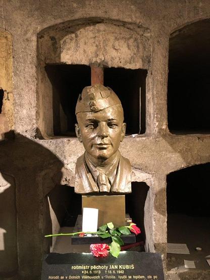 close-up of  bronze bust of assassin Jan Kubiš in garrison cap front of the burial cubicles

beneath each bust is a black plaque with names and dates of birth and death, rank, and biographical details in gold

visitors have placed a red rose, a red carnation, and other small tokens on the stand and plaque