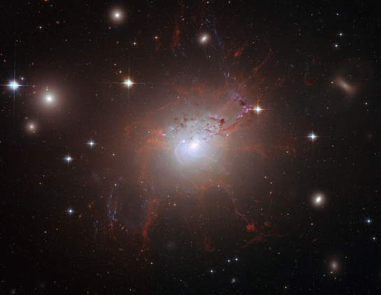 A Hubble image of galaxy NGC 1275, showing a bright central core surrounded by glowing red filaments of ionized gas. The filaments stretch outward in intricate, thread-like shapes against a dark background scattered with stars and distant galaxies.