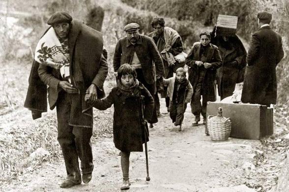 A group of individuals walking along a dirt path, carrying bundles and luggage. The scene depicts a somber atmosphere, with a mix of adults and children, including a girl with a cane. The environment appears rural and the photograph is in black and white.