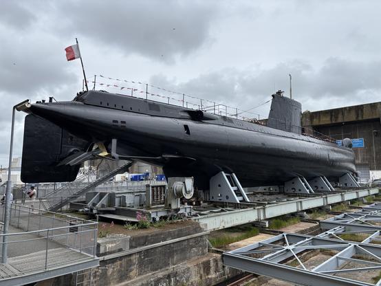 Černá vojenská ponorka vystavená na suchém molu s francouzskou vlajkou, umístěná na podpěrné konstrukci v přístavním areálu pod zataženou oblohou.