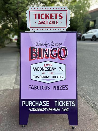 A very ornate A-frame advertising tickets for bingo, painted primarily in orchid purple, with an attachment shaped like a paper ticket affixed to the top.