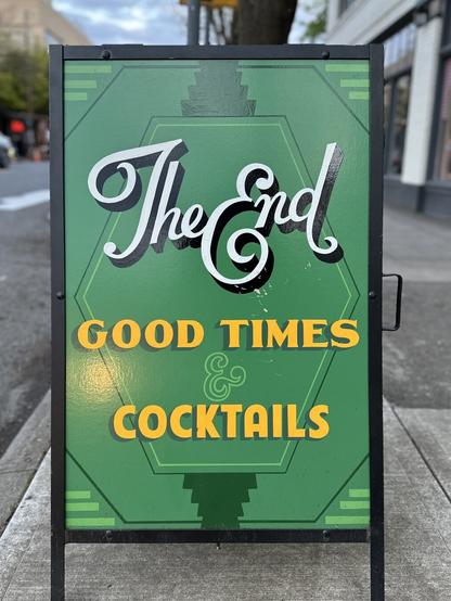 An A-frame advertising a bar attached to a movie theater, painted in emerald green and yellow. Lettering is reminiscent of old title cards.