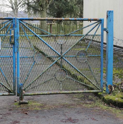 Rusty blue metal gate with intricate diamond and circle patterns, mounted on a concrete pathway. The gate leads to a fenced area with a moss-covered ground and various trees in the background. A building with a beige wall is partially visible on the right. The gate latch and hinges show signs of wear and age.