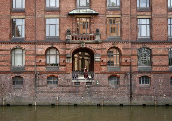 Photo d’une grande façade de vieil entrepôt en brique rouge dont le bas baigne dans une eau brun-vert. Au milieu, une ouverture laisse entrevoir une cour intérieure. On distingue une jeune femme debout contre le garde-corps fermant cette ouverture et un jeune homme assis par terre à côté. La façade est percée de nombreuses fenêtres et agrémentée de rayures en brique sombre.