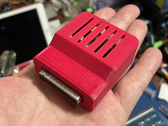 A little magenta box being held in a hand. It has a notch on one side near a 23-pin D-sub connector. There are some vents on the top.