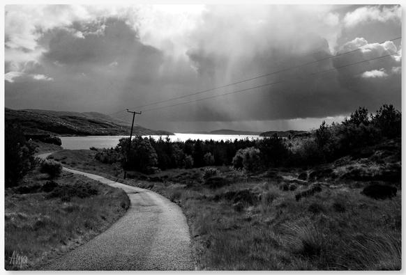 Eine einspurige Straße führt in einer Kurve auf einen See zu. Der Himmel ist dunkel und wolkenverhangen, einige Sonnenstrahlen scheinen durch und bringen den See zum Leuchten. Ein Strommast steht rechts am Straßenrand, Berge im Hintergrund. 

A single-lane road leads round a bend towards a lake. The sky is dark and cloudy, a few rays of sunshine shine through and make the lake glow. An electricity pylon stands on the right-hand side of the road, mountains in the background.