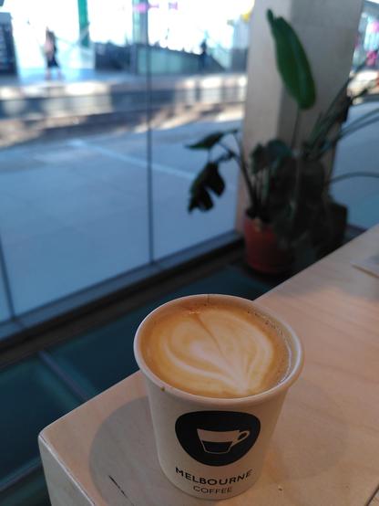 Close up of a small coffee drink on a wooden table, with train tracks and a large green plant in the background.