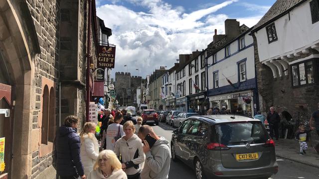 This picture captures a lively street scene in Conwy, Wales. The street is bustling with people walking along the sidewalks, some engaging in conversation or browsing items displayed outside shops. On the left side, there is a sign for the "Conwy Gift Shop" and another sign for a "Chip Shop." The buildings are a mix of stone and white-painted structures with black timber framing, reflecting traditional Welsh architecture.

Cars are parked along both sides of the narrow street, with a gray Vauxhall Zafira (license plate CX17 RLU) prominently visible. Flags, including the Welsh dragon, hang from some of the buildings. In the background, Conwy Castle with its distinct towers is visible, adding a historic charm to the scene under a partly cloudy sky with patches of blue. The atmosphere appears vibrant and busy, likely on a pleasant day.