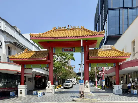 The image depicts a traditional Chinese-style archway, often referred to as a paifang or gate, characterized by ornate decorations and a vibrant color palette. The structure features a red and gold color scheme with intricate detailing on the roof, along with two lion statues on either side of the entrance. The background includes modern buildings, indicating that this archway is likely part of an urban area, possibly a Chinatown or a culturally themed district. The overall setting is bright with clear skies.

Image Credits: Wikimedia / Kgbo / CC BY-SA 4.0