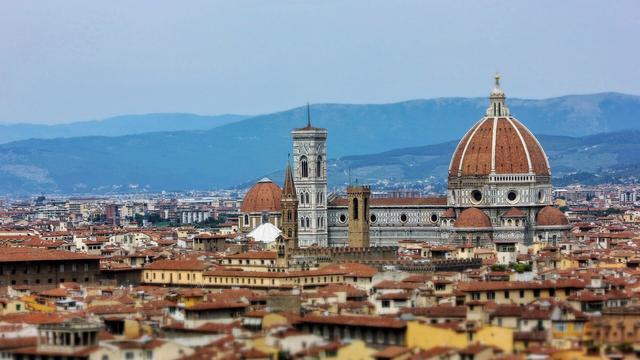 An elevated, slightly blurred view of the terracotta rooftops of Florence, Italy, with the prominent Florence Cathedral (Duomo) and its large red-tiled dome and Giotto's Bell Tower dominating the centre. In the background, blue mountains rise against a light grey sky, and the cityscape stretches out towards the horizon.