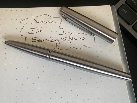 Una pluma metálica de formas limpias con el plumín carenado apoyada sobre un cuaderno en el que se ha escrito “Jueves de estilográficas” en tinta marrón