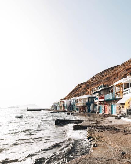 Colorful seaside houses built into a rocky hillside line the edge of the water, with gentle waves washing up against the shore. The sky is bright and clear, and a small boat floats in the sea near the houses.