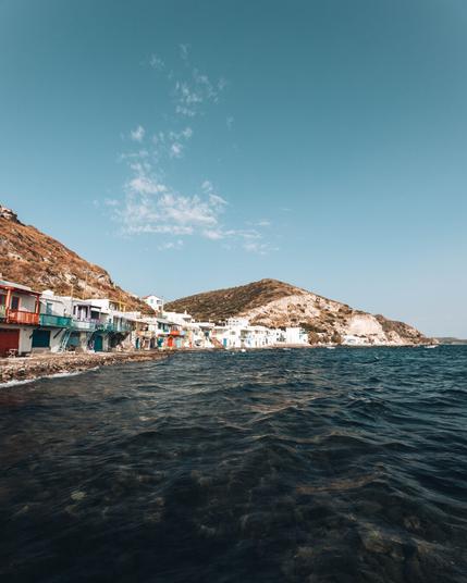 A row of colorful houses with balconies lines the rocky shoreline of a coastal village, with clear blue water in the foreground and hills rising in the background under a bright, mostly clear sky.