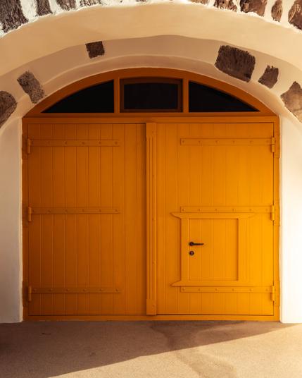 A large, bright yellow double wooden door with a small inset door in the right panel, set in a white arched wall with stone accents. Sunlight casts shadows on the ground in front of the door.