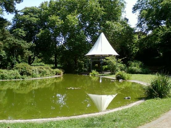 Ein Teich in einem Park. Hinten rechts ein Pavilion mit weissem Dach.