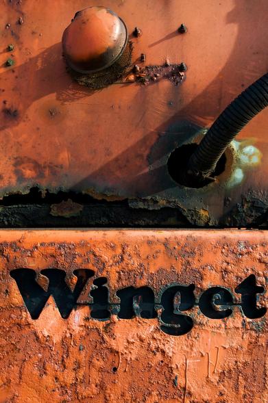 Close-up of an orange, weathered surface featuring rust and wear, with the word "Winget" prominently displayed in black lettering. A rounded metal component is also visible.