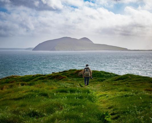 A man with a camera walking towards the edge of a green grassy cliff with the ocean and an island in the distance and cloudy skies above.
