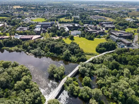 A drone photograph of the University of Limerick campus. There is a wide river lined with dense, mature trees. The river is spanned by a long, curved pedestrian bridge. On the far side of the river there is a sprawling lawn surrounded by trees and many buildings nestled amongst them
