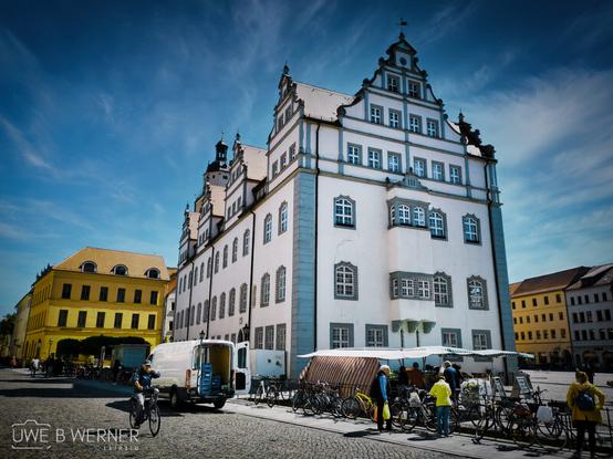 Das Bild zeigt das historische Rathaus von Wittenberg mit seiner prächtigen Renaissancefassade. Vor dem Gebäude befinden sich Marktstände, Fahrräder und Passanten bei sonnigem Wetter auf dem Marktplatz.