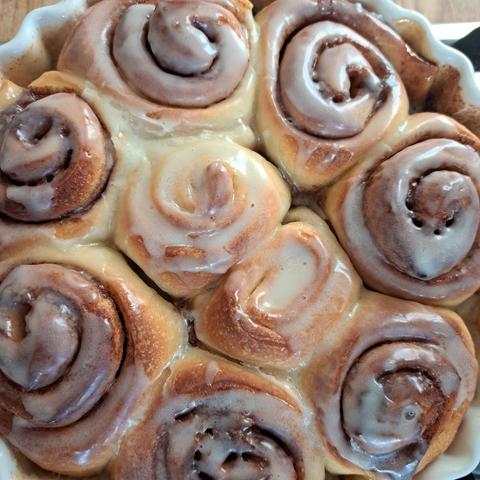 A round ceramic flan dish containing freshly baked sourdough cinnamon rolls. The rolls are nestled together snugly. The centre two are a paler golden colour. The icing glaze is pooled in the centre of one of the two middle ones.