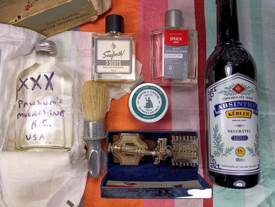 (Le Chat Mistral description)

The image displays a collection of grooming and personal care items arranged on a striped cloth. On the left, there is a clear bottle labelled "Pausum's Moonshine, N.C., USA" with purple XXX markings. Next to it, there's a shaving brush with a metallic handle. In the centre, there's a small bottle of Seaforth! 3 Scots Eau de Toilette and a tin of Martin de Candre shaving soap. Above these, there's a bottle of Speick Men Active After Shave Lotion. To the right, there's a bottle of Swiss Absinthe Kübler. At the bottom, there's an open box containing a vintage-style metal tool. The items are neatly arranged, creating a visually appealing display.