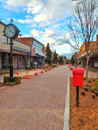 The pedestrianised main street of Windsor. The middle of the street is paved with bricks. There are milch garden beds with trees on both the left and right. The street is lined with historic buildings. There is an old clock and a red latter box ahead.