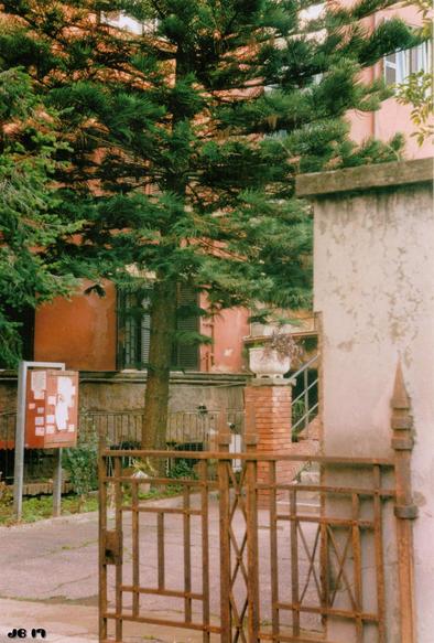 Farbfoto, das den Eingang zu einem Wohngebäude-Innenhof in Garbatella, einem Stadtteil Roms mit Bauten aus den 1930er-Jahren zeigt. Im Vordergrund eine rostige Eisenkonstruktion, die wohl in der Vergangenheit den Zugang beschränkt hat. Im Bildzentrum ein großer Nadelbaum. Leica IIIg mit 50 mm Summitar auf Kodak-Film.