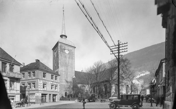 Black and white photograph of Korskirken in Bergen, Norway from the 1920s. The church features a tall steeple with a clock on its tower. Surrounding buildings include shops with signs, one reading "Simonsen & Co." Vintage cars and horse-drawn carriages are visible on the street, along with pedestrians. Power lines stretch across the sky.