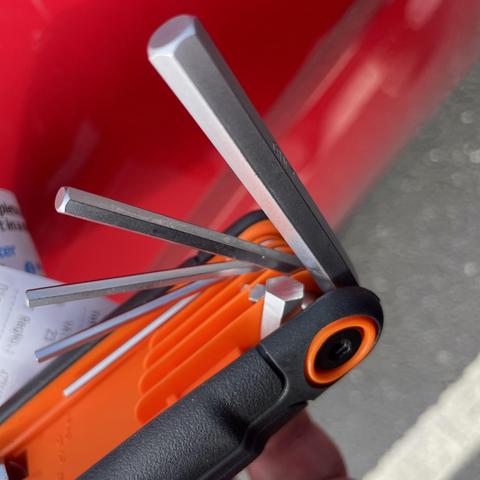 An assortment of hex keys stored in an orange and black holder, with several keys partially extended. A red background is visible, along with some blurred objects in the background.