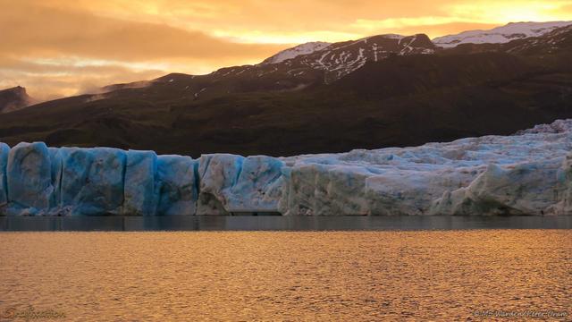 A colour photo of a body of water at sunset, with the slightly ruffled water reflecting the orange and yellow of the sky. On the horizon is a mountain face topped with snow. A band of cyan ice is standing at the water's edge, deeply cracked and crevassed.