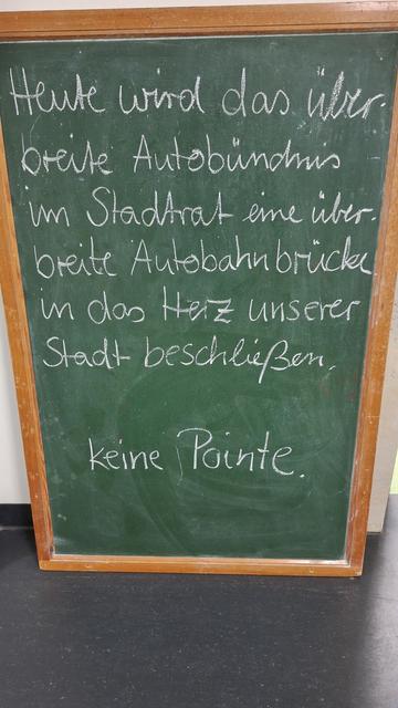 Auf einer grünen Tafel steht mit weißer Kreide geschrieben:
Heute wird das überbreite Autobündnis im Stadtrat eine überbreite Autobahnbrücke in das Herz unser Stadt beschließen. Keine Pointe.