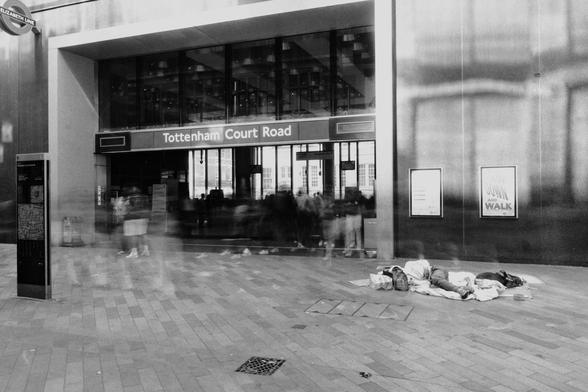 Long exposure photo of an entrance to a tube station.
Blurred shapes of people in hurry to get somewhere and a still body of a homeless person and their dog.