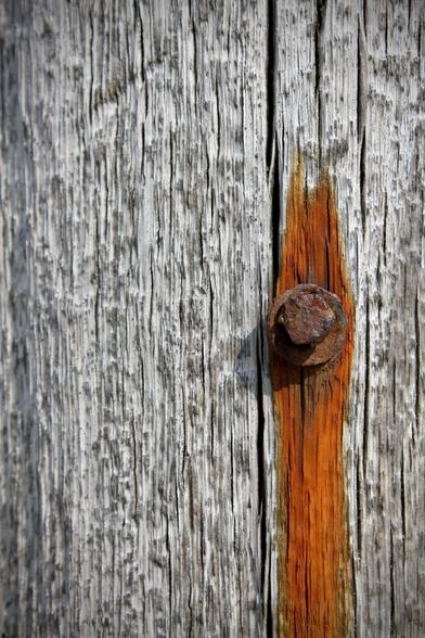 das bild im hochformat zeigt einen verwitterten holzpfosten aus grauem, rissigem holz. am rechten bildrand auf mittiger höhe steckt ein rostiger bolzen im pfosten, der von einer vertikal verlaufenden, orangefarbenen rostspur umgeben ist.
der kontrast zwischen dem vergrauten holz und dem leuchtend orangenen rost fällt stark ins auge. die oberfläche des holzes wirkt trocken und spröde.