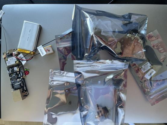 An assortment of electronic components laid out on a metallic surface. Items include a Raspberry Pi board, a battery, a GPS module, and several sealed bags containing circuit boards and connectors.
