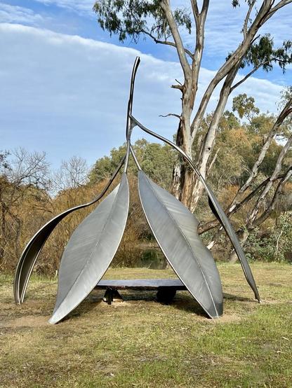 Four large metal guy leaves looking like they’ve just fallen from a tree, with a raised platform inside. A eucalyptus tree is framed behind it & native bush behind that. All framed against a lovely cloud filled sky with a bright streak of blue near the top.