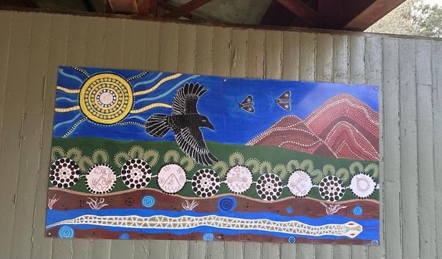 I don’t know if I’ll do this justice, but I’ll try. 
Indigenous art work on a pylon under an old railway bridge. It shows a yellow sun in the corner with rays streaming out, beside it is a black bird, like a raven plus some Bogong moths in the sky, possibly food for the raven. Then beside that are some red tinged hills. Beneath are representations of grasses, bushes, plants & flowers. Beneath again is the soil with plant root systems, then what looks like water with a patterned serpent swimming through.