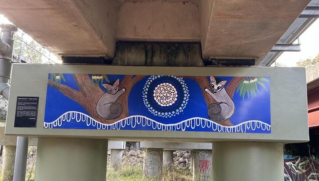 Between two old railway bridge pylons with indigenous artwork of two possums sitting in the forks of trees on either side of the picture. In the middle is what I believe may be the moon. I’m not sure what the white wavy lines represent but for me I see them fluid, like dew.