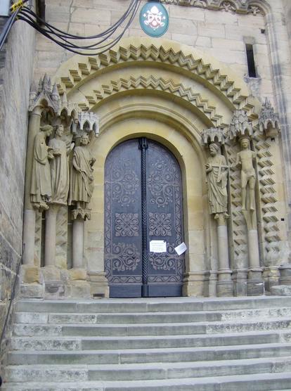 Tall wooden double-doors recessed in a stone wall. There are two concentric arches with zigzag designs above it and three statues of human figures to the left, and two statues of human figures to the right.