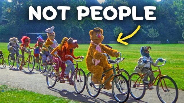 A group of Muppet characters including Fozzie Bear ride bicycles through a green park setting with "NOT PEOPLE" text and a yellow arrow pointing to demonstrate that no human puppeteers are visible in the outdoor scene.