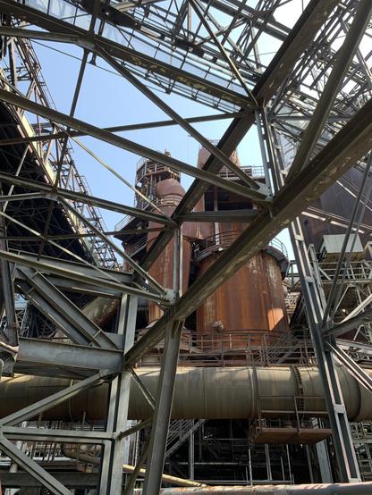 Aufnahme von zwei metallenen zylinderförmigen Industriebauten, die von außen verrostet sind. Sie befinden sich in der Mitte des Photos. Die Ansicht wird zum Teil durch kreuz und quer im Bild verlaufende Metallverstrebungen und Stützen versperrt. Im Hintergrund hellblauer wolkenloser Himmel.

Photo of two metal cylindrical industrial buildings that are rusted on the outside. They are in the center of the photo. The view is partially obstructed by metal struts and supports running criss-cross through the picture. In the background is a light blue, cloudless sky.