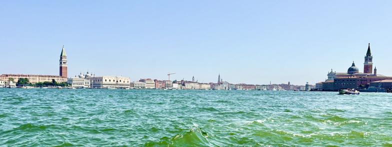 This image shows a wide, panoramic view of Venice seen from the water. In the foreground, greenish-blue waves of the Venetian lagoon shimmer under a clear blue sky. On the left side of the image stands the tall Campanile of St. Mark’s Basilica, a brick bell tower with a green pointed roof. Next to it are the white domes of St. Mark’s Basilica and the ornate white facade of the Doge’s Palace. 

Across the center of the image stretches the historic cityscape with pastel-colored buildings. On the right side of the image is the island of San Giorgio Maggiore with its large church dome and a red brick bell tower, both clearly visible. Small boats float on the water, adding to the maritime atmosphere.