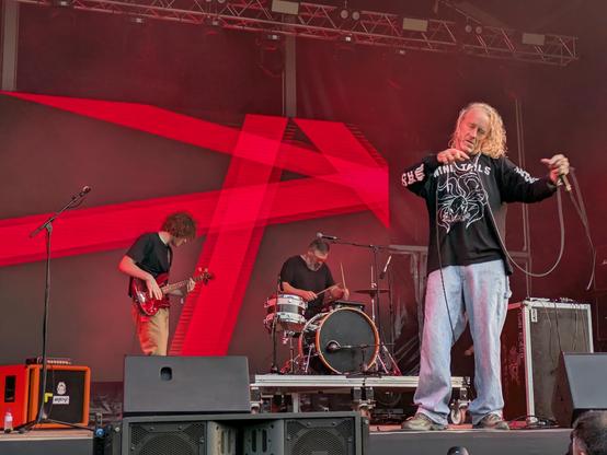 Three musicians on stage against a dark background with bold red stripes in various directions. Left to right, a bassist and drummer on a minimal kit in the middle distance. The singer has a NineTails sweatshite, long white-blond straggly hair and is lost in his musical world. It is Alan Sparhawk with his band