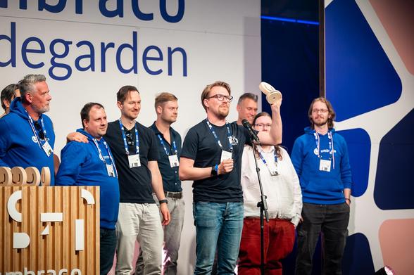 A group of team members stand on stage at an event. One team member, standing at a microphone, holds up a wooden Umbraco award while giving a speech. Others around him smile and applaud. Everyone is wearing event lanyards, and some are dressed in branded Umbraco hoodies. The stage backdrop reads “Umbraco” and “Codegarden.”